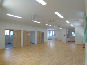 児童クラブの内観1。広々としたホール。室内は段差が少なくバリアフリーに配慮された造り。女子トイレ・多目的トイレ・男子トイレがあり、明るい色調の引き戸で仕切られている。