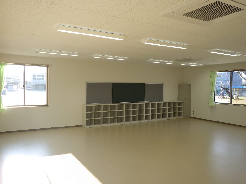 児童クラブの内観1。左斜めから。広々とした室内の後部に黒板と収納棚が配置されている。