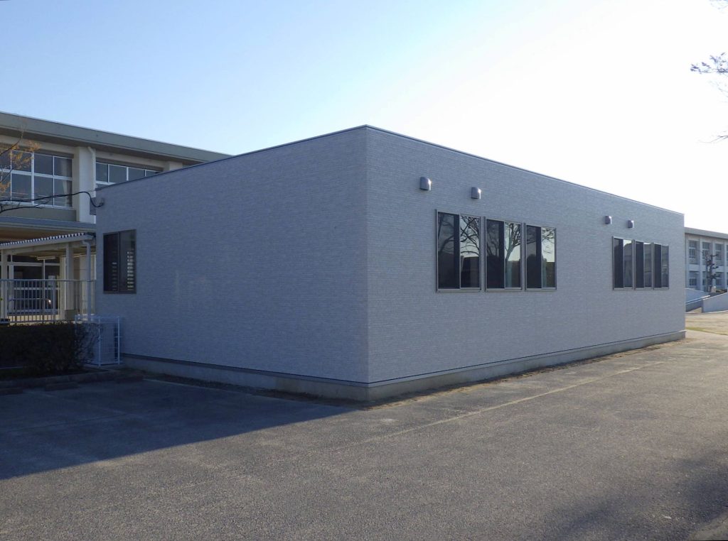 児童クラブの外観1。建物裏側斜めからの全景。ベージュの壁に窓と排気フードが取り付けられている。