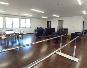 デイサービス施設の内観1。談話室。広い室内にテーブルセットやソファで歓談スペースが設けられており、手前には機能訓練のための平行棒も配置されている。
