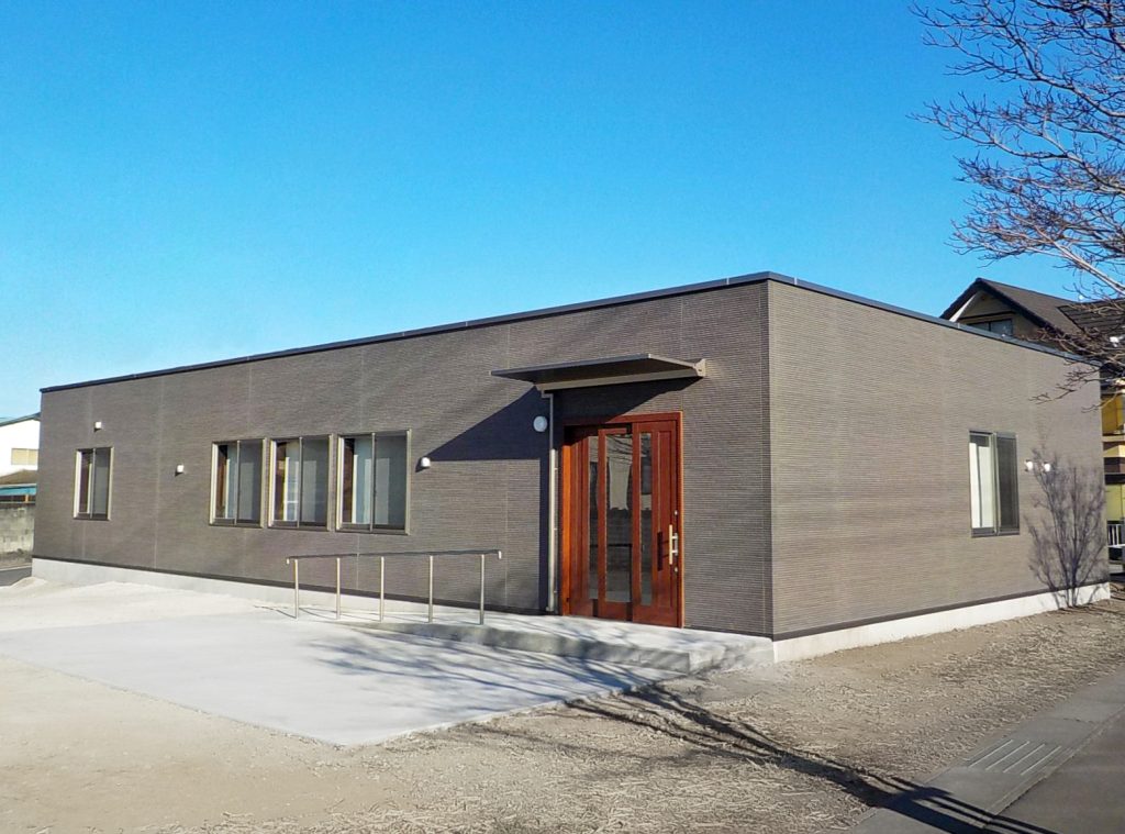 デイサービス施設の外観1。右斜めからの全景。緩やかなスロープと手すりが配された広めの引き戸の玄関の様子。