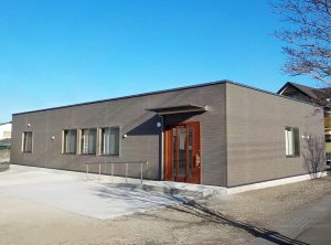 デイサービス施設の外観1。右斜めからの全景。緩やかなスロープと手すりが配された広めの引き戸の玄関の様子。